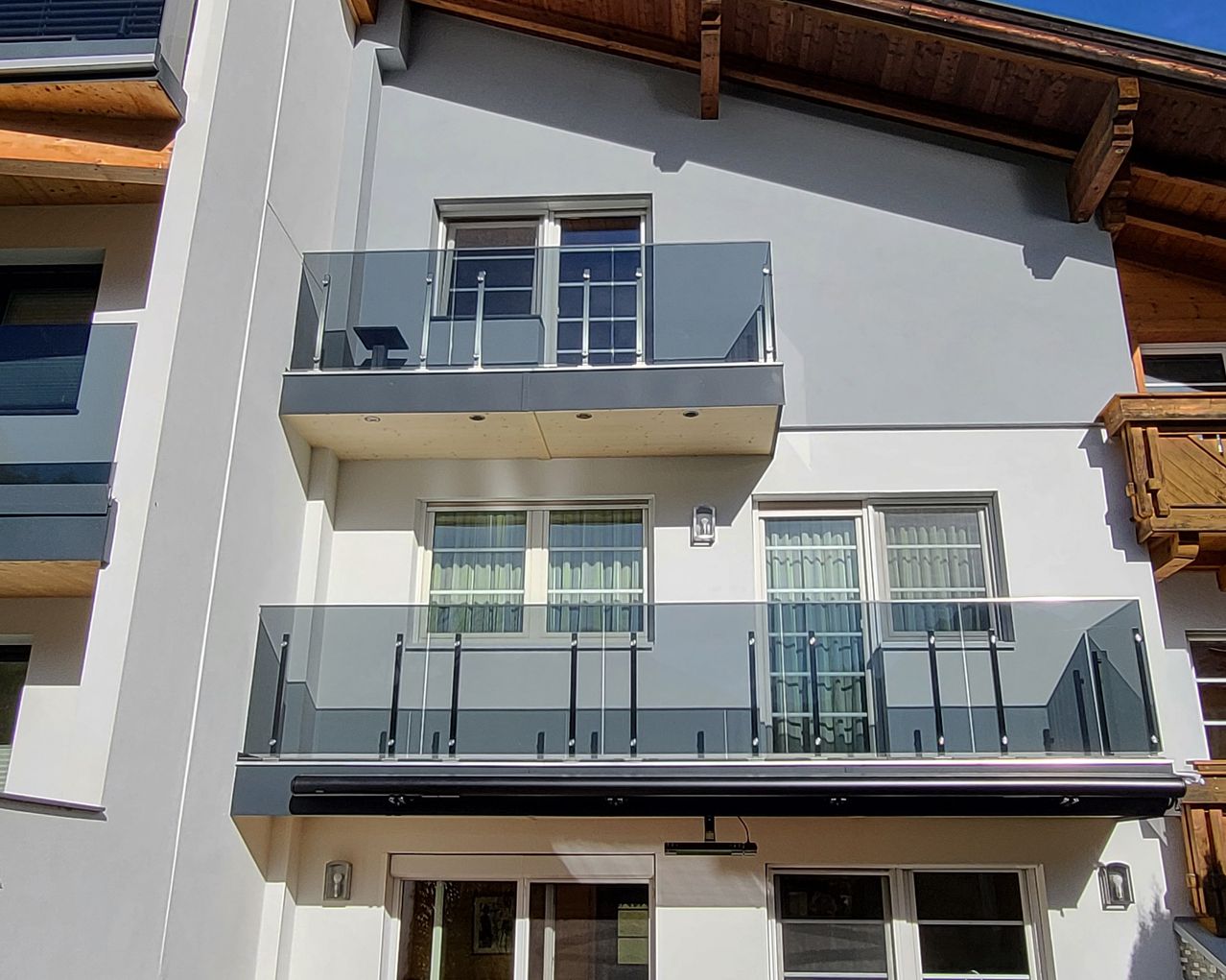 Ein modernes zweistöckiges Haus mit Glasbalkonen und Fenstern. Die Balkone haben Glasgeländer und die Fenster Vorhänge. An der Wand neben dem Eingang befindet sich eine Lampe.