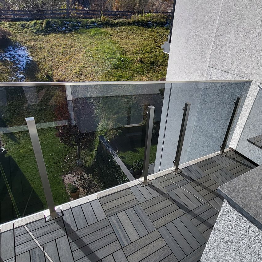 Ein Balkon mit einer Glasbrüstung und Blick auf ein Grasfeld und ein Gebäude. Der Balkon hat einen Holzboden.