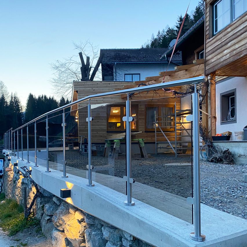 Ein modernes Haus mit Holzfassaden, einem neuen Glasgeländer an der Seite und einem Schotterbereich. Bäume und ein weiteres Haus im Hintergrund.