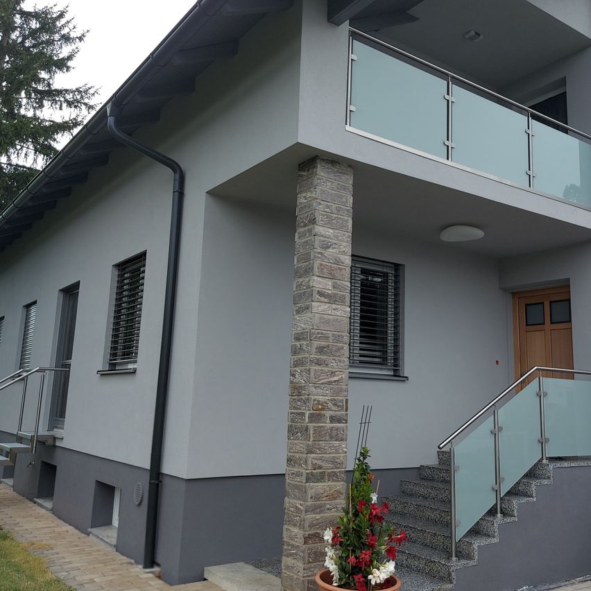 Ein modernes Haus mit einem Balkon mit Glasgeländer, grauen Wänden und einer Holztür. Ein kleiner Topf mit einer Pflanze steht vor den Stufen.