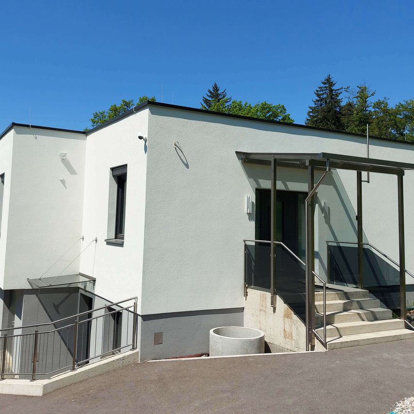 Ein modernes weißes Gebäude mit einem grauen Dach, Glasfenstern und Metallgeländern an den Treppen und der Veranda. Im Hintergrund sind Bäume sichtbar.