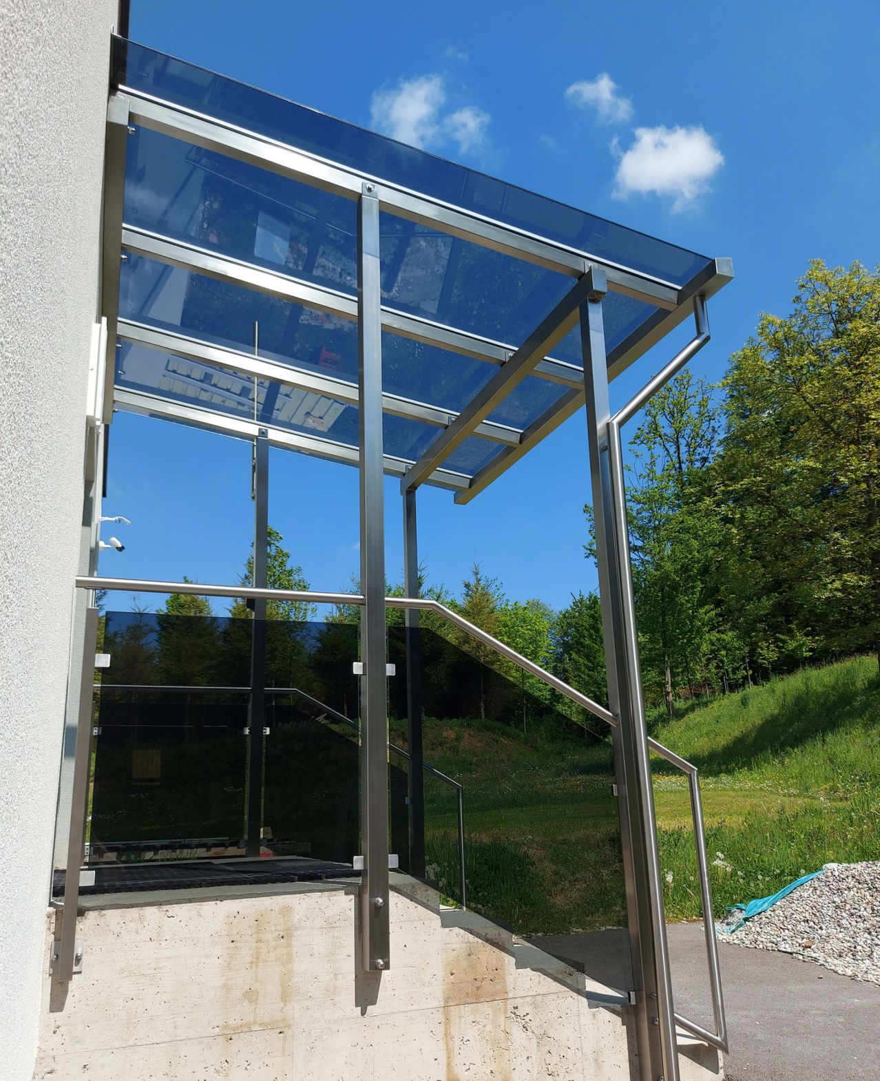 Ein moderner Eingang mit Glasgeländern und einem Stahldach befindet sich neben einem Grashügel unter einem klaren blauen Himmel.