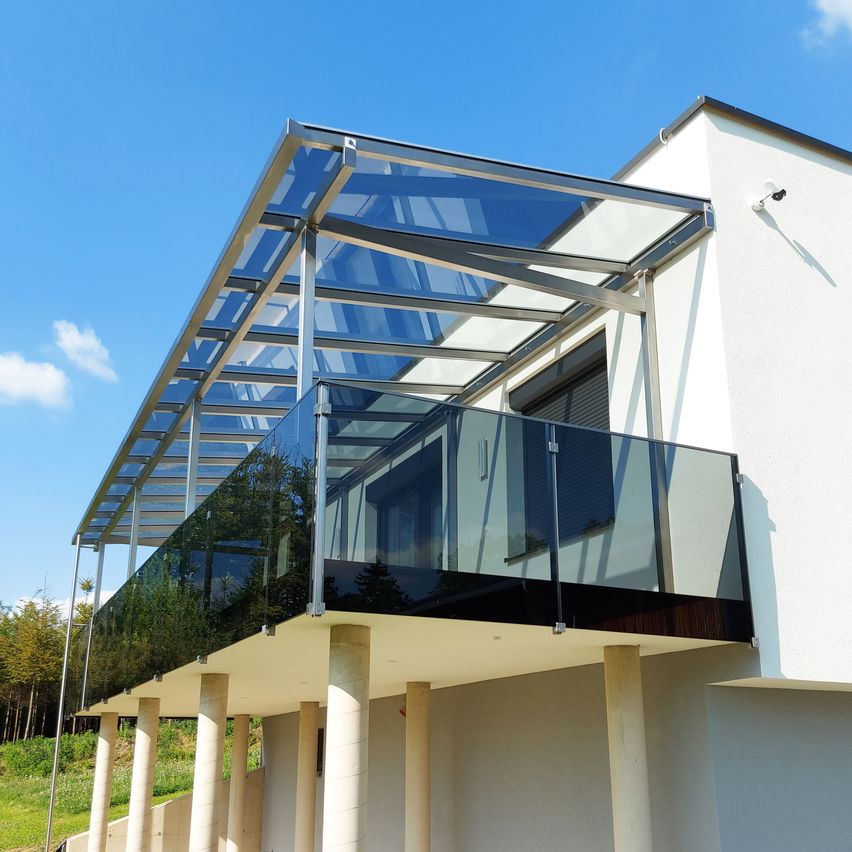 Ein moderner Balkon mit einer Glasbrüstung und einem transparenten Dach ist an einem weißen Gebäude unter einem klaren blauen Himmel befestigt.