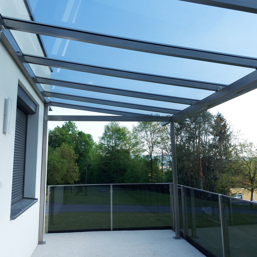 Ein Balkon mit Glasdach und transparenten Geländern blickt auf eine Rasenfläche und Bäume. Der Himmel ist blau mit einigen Wolken.