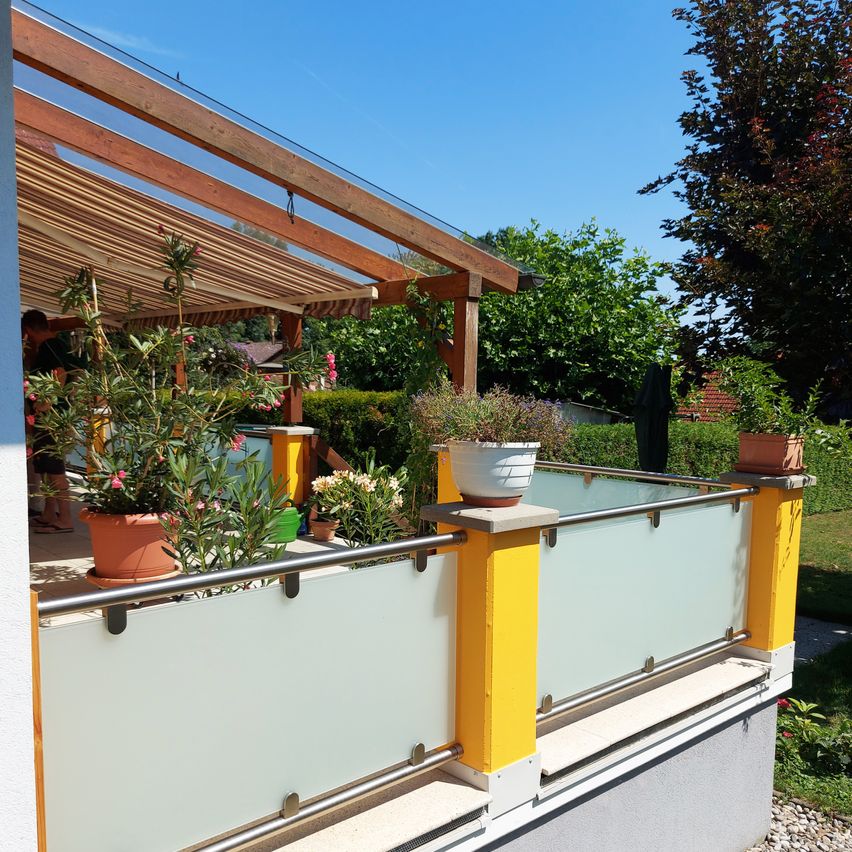 Ein Balkon mit Glasgeländer und Topfpflanzen, eine Holzpergola und Blick auf einen sonnigen Tag mit Bäumen im Hintergrund.