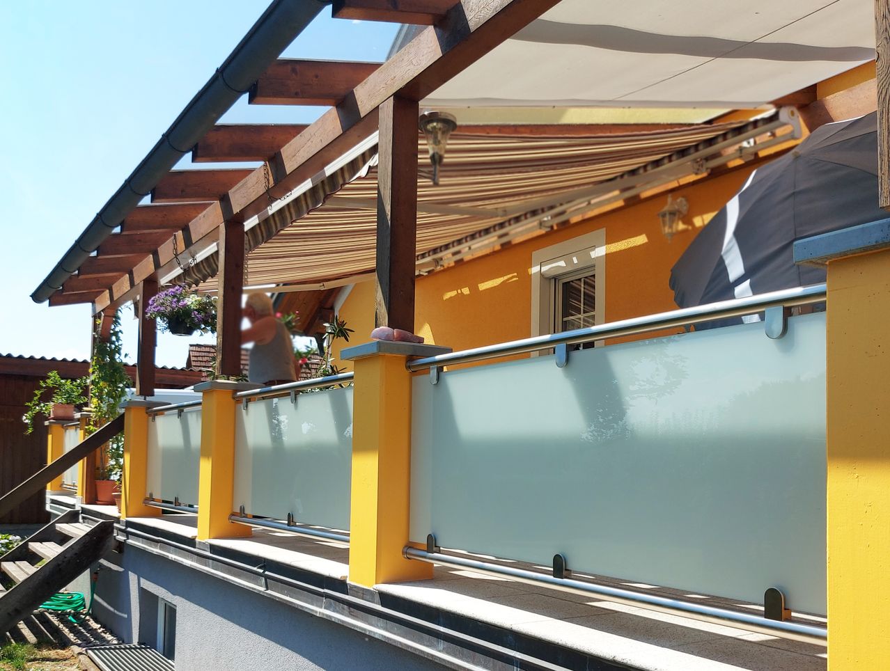 Ein Balkon mit gelben Geländern und einer Glasabtrennung, beschattet von einem versenkbaren Markisen, befestigt an einem gelben Gebäude mit einem weißen Fenster und einer hängenden Pflanze.