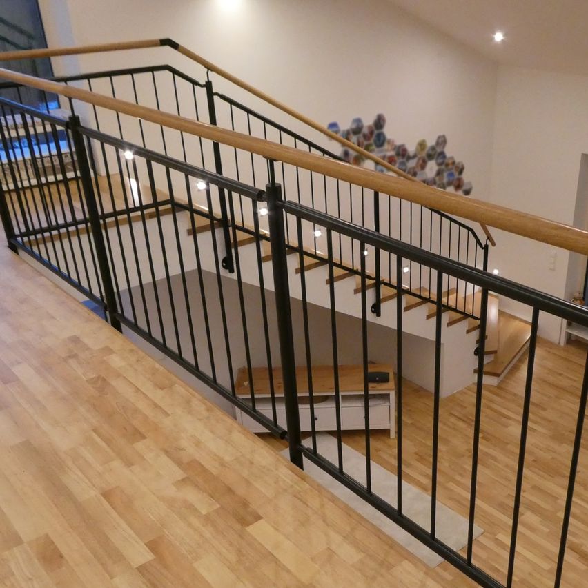 Eine Holztreppe mit schwarzen Geländern und Holzhandläufen ist zu sehen. Die Treppe führt in eine obere Etage. Der Holzboden ist unten sichtbar. Eine weiße Wand befindet sich an der Seite.