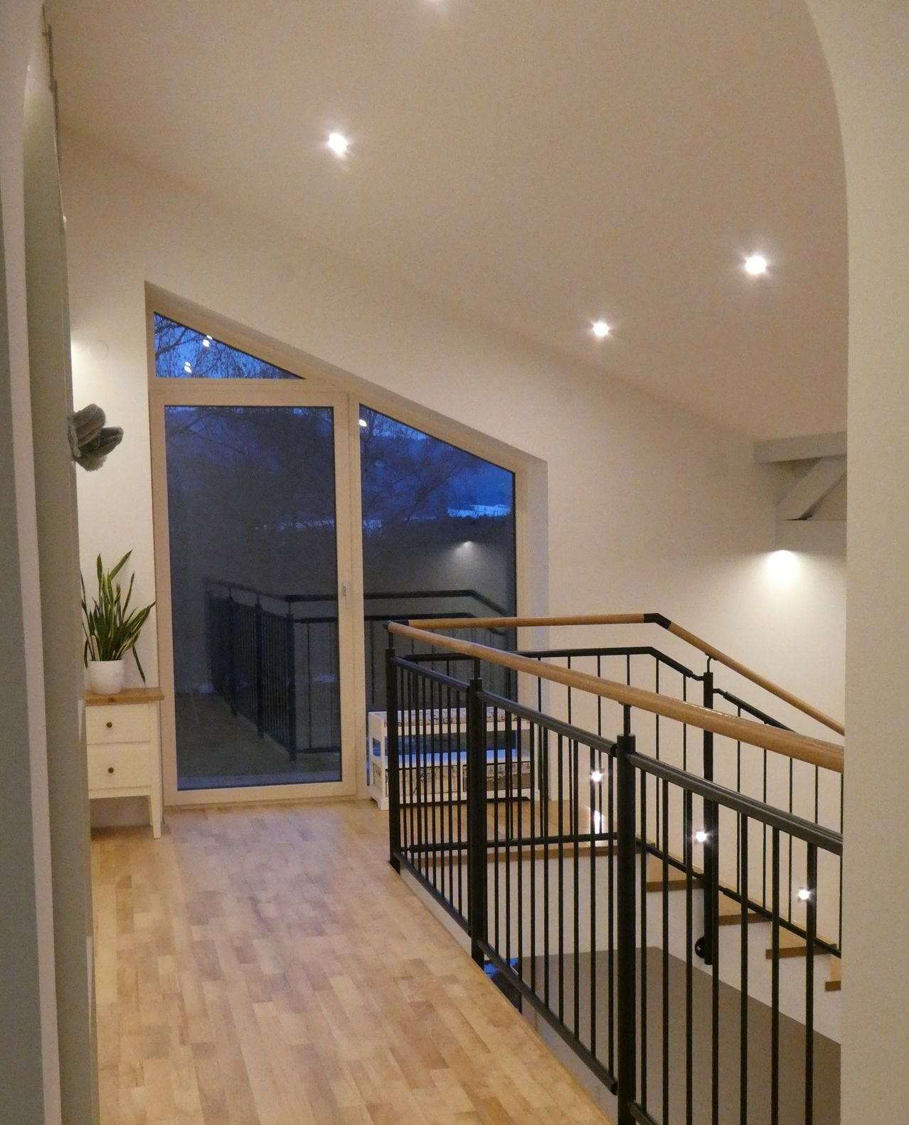 Ein modernes Interieur mit Holzboden, weißer Wand und einer Treppe mit schwarzem Geländer. Es gibt eine Glastür mit Balkon im Freien.