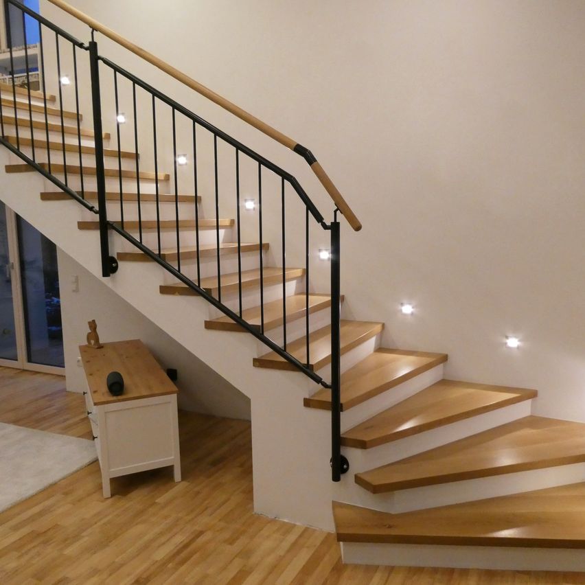 Eine moderne Treppe mit Holzstufen und schwarzen Geländern. Die Treppe hat an jeder Stufe ein Licht. Links befindet sich ein weißer Holztisch mit einem schwarzen Objekt obenauf.