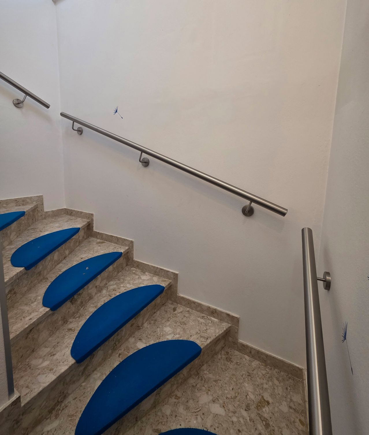 Eine weiße Marmortreppe mit blauen Handläufen und blauen Stufen. Die Wand ist weiß gestrichen mit einem kleinen Löwenzahn-Aufkleber.
