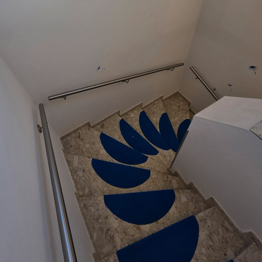 Eine Treppe mit weißen Wänden und Marmorstufen ist zu sehen. Blaue Matten sind auf den Stufen platziert, und Metallgeländer sind auf beiden Seiten installiert. Die Wände sind mit Löwenzahn-Designs verziert.