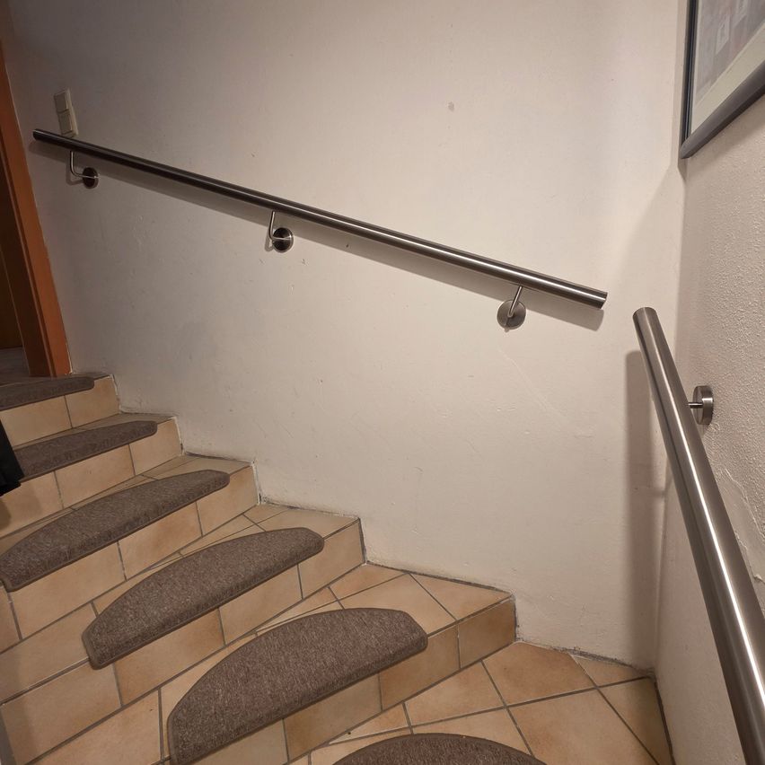 Das Bild zeigt eine Treppe mit einer weißen Wand, einem Metallgeländer und gefliesten Stufen mit grauem Teppich.