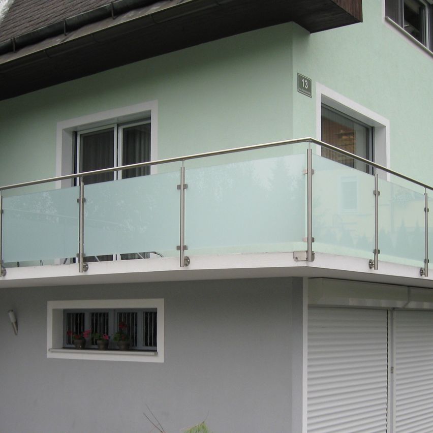 Ein Haus mit Balkon, Glasgeländern und einer Garage. Das Haus hat grüne Wände, weiße Fensterrahmen und die Nummer 13 an der Wand. Der Balkon hat ein Fenster mit Topfpflanzen.