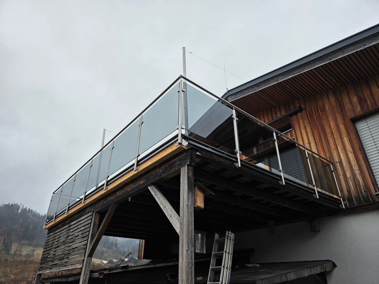 Ein Balkon mit Glasgeländern, befestigt an einer Holzkonstruktion unter einem bewölkten Himmel.