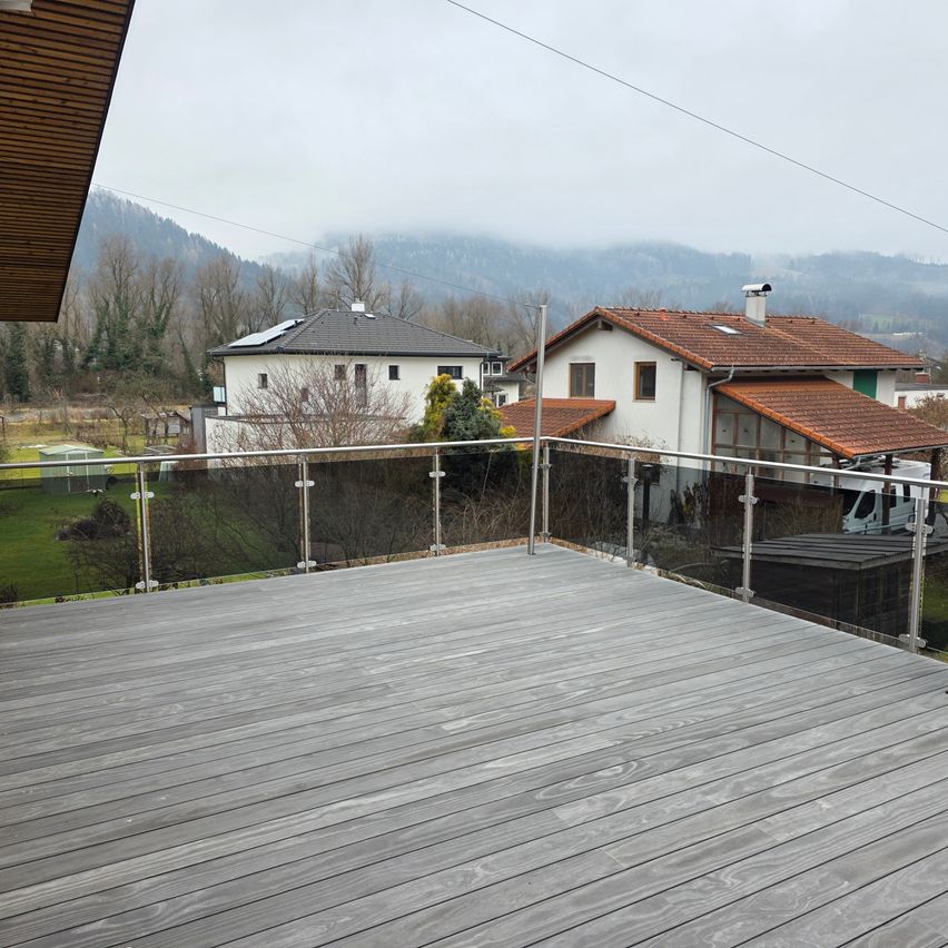Ein Balkon mit Metallgeländer befindet sich vor zwei Häusern. Beide Häuser haben Solarmodule auf den Dächern und sind von Grün umgeben. Der Balkon ist leer, und der Boden ist mit Holz bedeckt. In der Ferne gibt es Berge und Bäume.