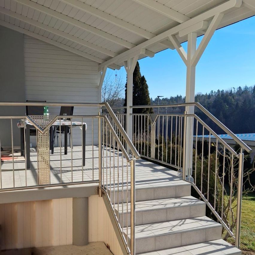 Eine Veranda mit Tisch und Stühlen ist an einem Haus mit weißen Wänden und einem weißen Dach befestigt. Die Veranda hat ein Metallgeländer mit Rautenmuster. Das Haus ist von Bäumen und einem blauen Dach umgeben.