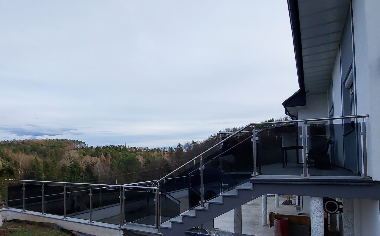 Ein Balkon mit Glasgeländern und Treppen, die zu einem ebenerdig gelegenen Bereich mit Bäumen und einem klaren Himmel im Hintergrund führen.