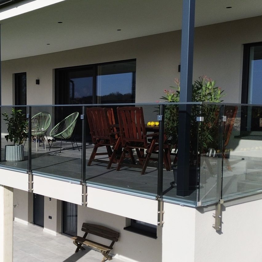 Ein Balkon mit Glasgeländer, mit Tisch und Stühlen, umgeben von Pflanzen, und einer Holzbank auf einem gefliesten Boden.