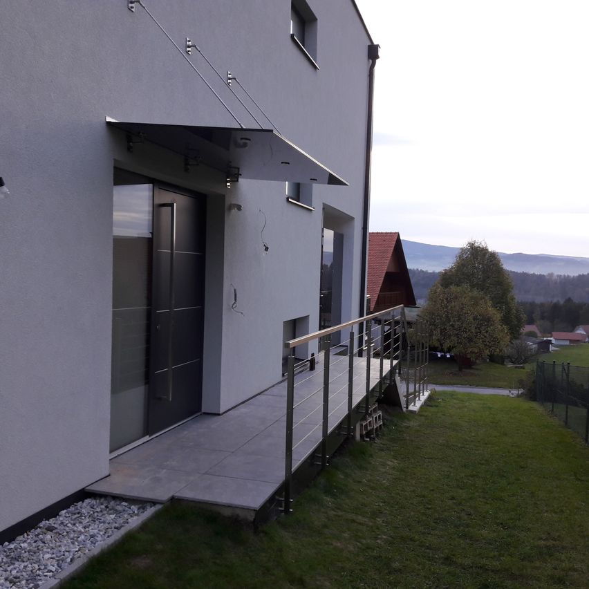 Ein graues Haus mit Balkon und Metallgeländern. Ein kleiner Garten mit Rasen und einem Baum im Hintergrund.