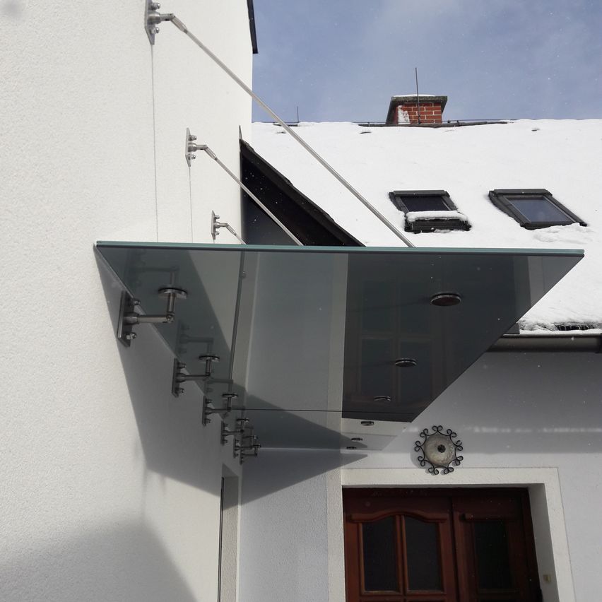 Eine moderne Glasüberdachung, die an ein weißes Gebäude angebracht ist. Sie erstreckt sich über den Eingang mit einem dekorativen Element über der Tür. Schnee bedeckt das Dach darüber.