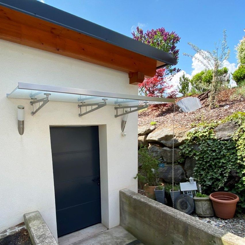 Ein modernes Haus-Außendesign mit einem Glasvordach über der Garagentür. In der Nähe befinden sich verschiedene Topfpflanzen und ein dekoratives Haus auf dem Boden.