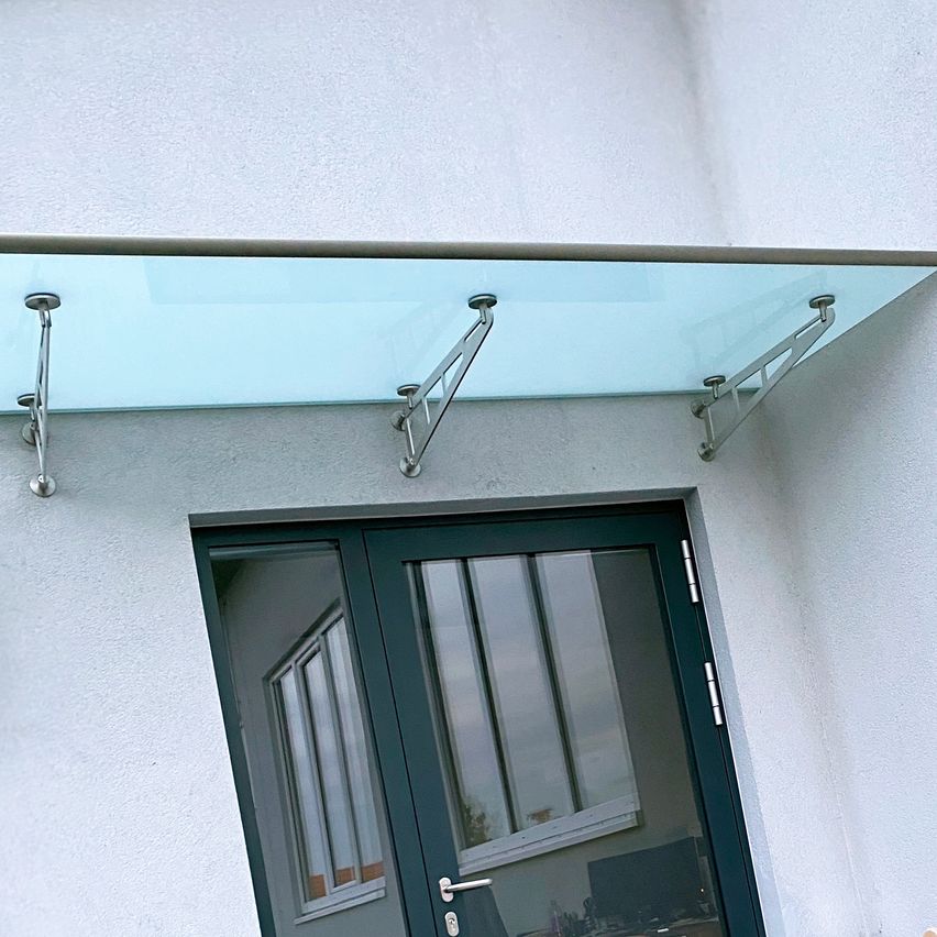 Ein Glasvordach mit Metallhalterungen ist über einem weiß verputzten Gebäude montiert. Darunter ist eine schwarze Tür mit Glasscheiben und einem silbernen Griff zu sehen.