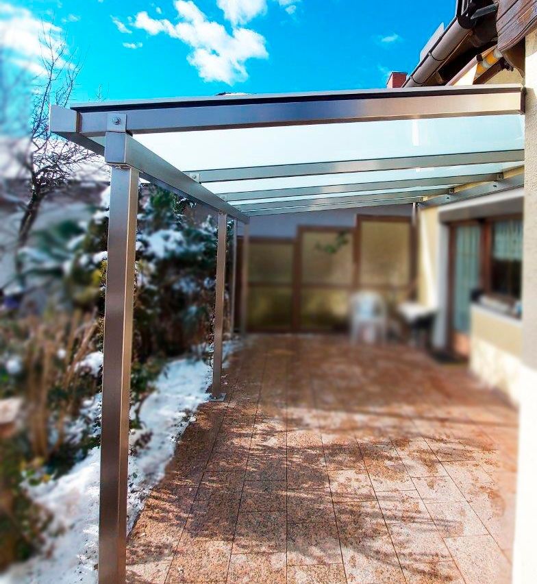 Eine Terrasse mit einem Glasdach, das von Metallpfosten getragen wird. Schnee liegt auf dem Boden, und ein Stuhl steht in der Nähe des Gebäudes.