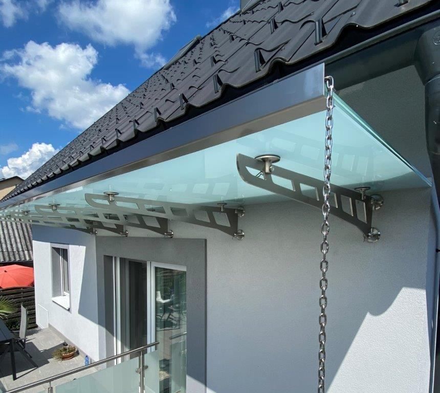 Ein modernes Haus mit einem Glasvordach, das von Ketten gehalten wird, unter einem teilweise bewölkten Himmel. Das Vordach ist am Dach befestigt.