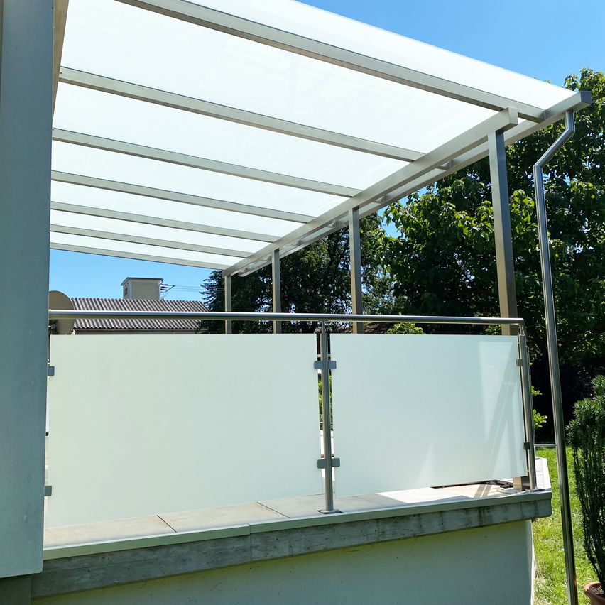 Ein Balkon mit einem Glasdach und Metallgeländern steht unter einem klaren blauen Himmel, umgeben von Bäumen und einem Gebäude im Hintergrund.
