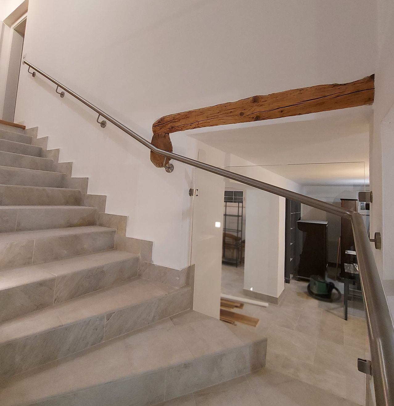 Eine weiße Treppe mit Marmorstufen führt zu einem Flur mit einer Glastür. Die Treppe hat einen Holzbalken über sich und eine Metallhandlauf ist an der Wand befestigt.
