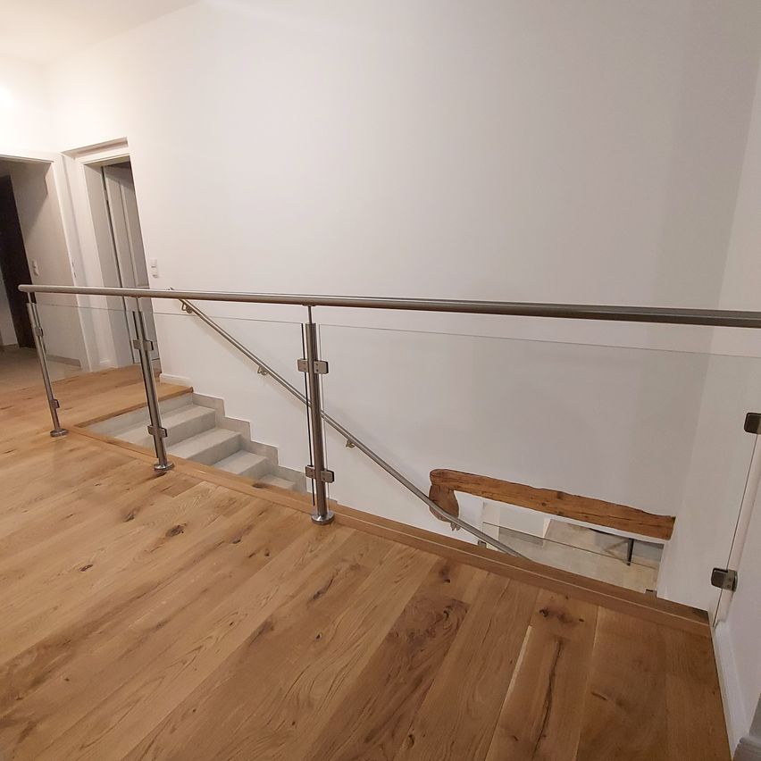 Eine moderne Treppe mit einer Glasbrüstung und Holzstufen. Das Geländer hat ein schlankes Metalldesign und eine Glasscheibe. Der Boden ist aus Holz und poliert. Die Wand ist weiß.