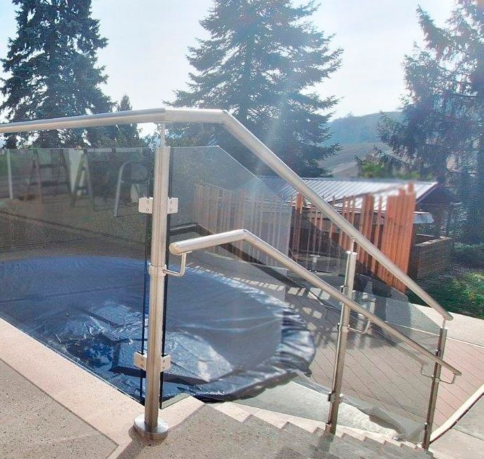 Eine Terrasse mit einer Glasbrüstung, einem blauen Tarp, das einen Pool bedeckt, und einem Holzzaun dahinter. Bäume und ein Gebäude sind im Hintergrund.