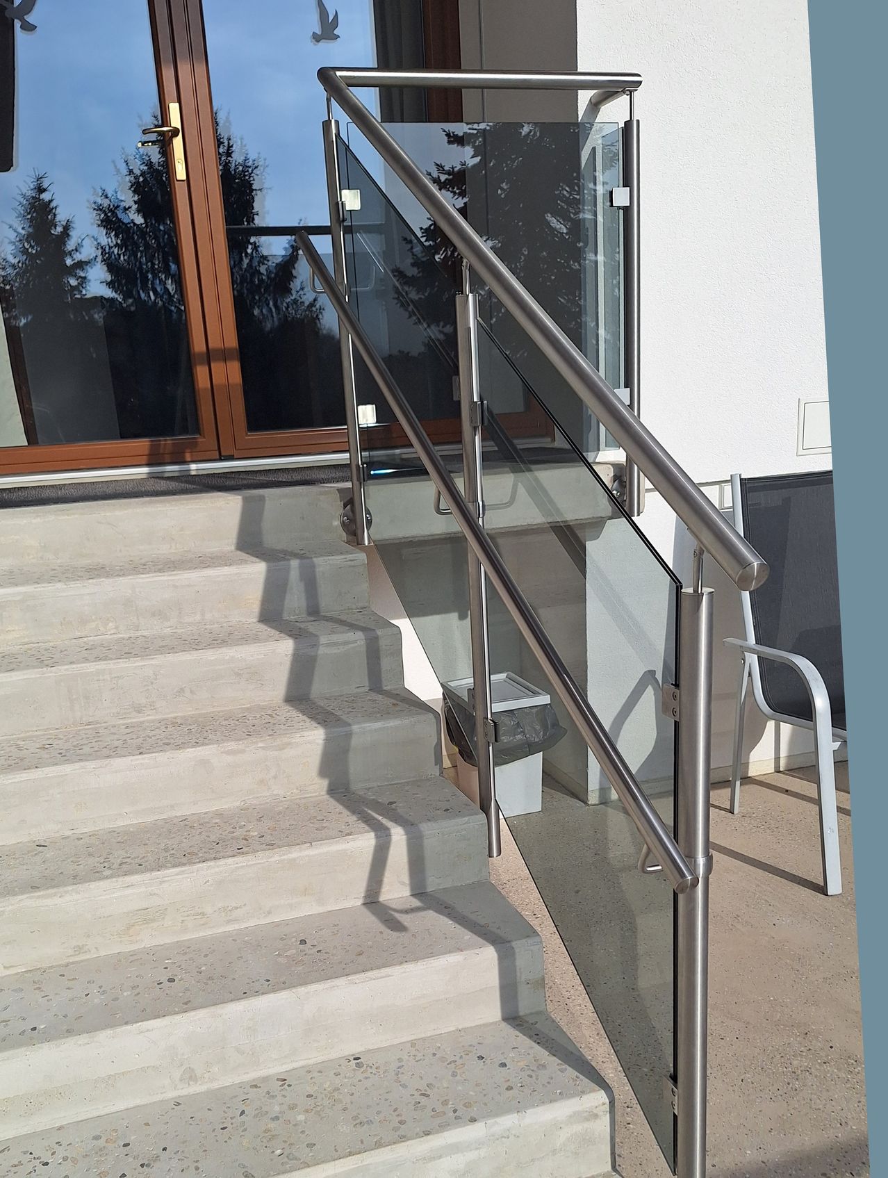 Ein moderner Hauseingang mit Glasstufen und einem Handlauf aus Edelstahl. Ein Mülleimer steht neben den Stufen. Bäume sind durch die Glastür sichtbar.