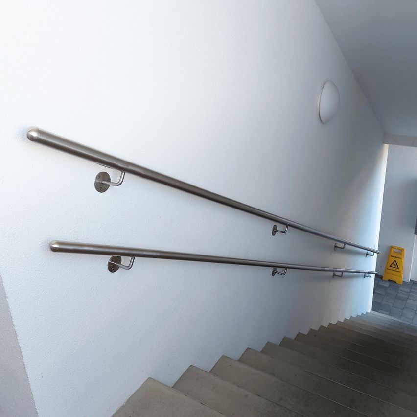 Eine weiße Wand mit zwei Edelstahl-Handläufen auf beiden Seiten einer Treppe, ein gelbes Warnzeichen befindet sich unten rechts.