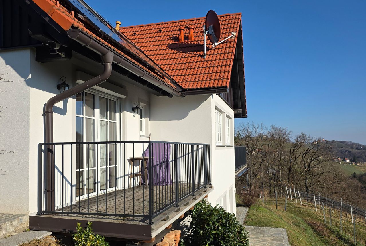 Ein weißes Haus mit rotem Ziegeldach und einer Satellitenschüssel ist zu sehen. Das Haus hat einen Balkon mit schwarzen Geländern. Im Vordergrund ist ein Tisch mit einem lila Bezug zu sehen.