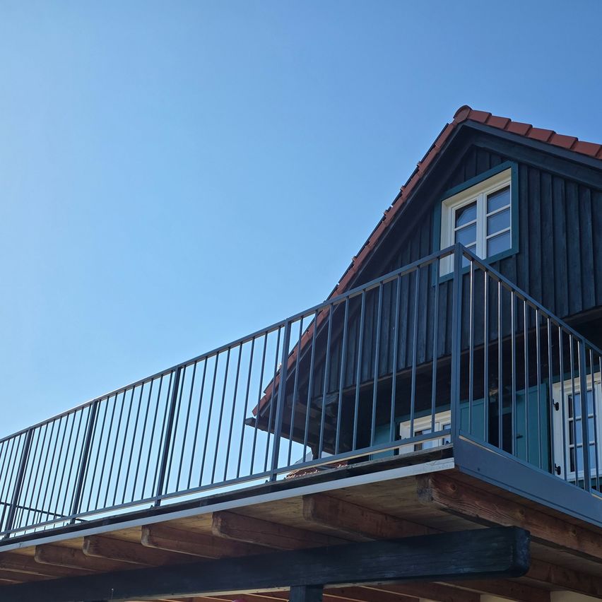 Ein hölzerner Balkon mit einer Metallbrüstung ist an der Seite eines Hauses befestigt. Das Haus hat ein braunes Dach und schwarze Wände.