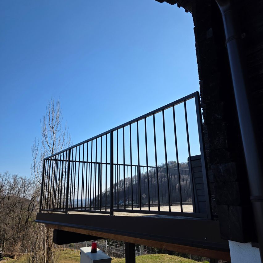 Ein Balkon mit einem schwarzen Metallgeländer und Blick auf einen Hügel mit Bäumen und einem klaren blauen Himmel.