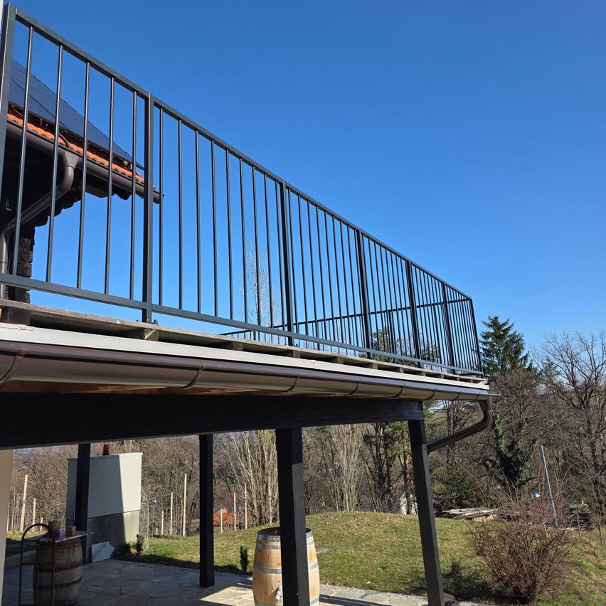 Ein Balkon mit schwarzen Geländern und Metallstützen ist an einem Haus unter einem klaren blauen Himmel befestigt, umgeben von Bäumen und Gras.