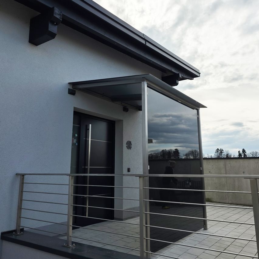 Ein moderner Hauseingang mit einer Glasüberdachung, schwarzer Tür und einem Metallgeländer auf dem Balkon. Der Himmel ist bewölkt.