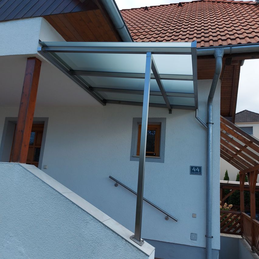 Ein modernes Haus mit einem Glasvordach über dem Eingang, Holzstützen und der Nummer 44 an der Wand.