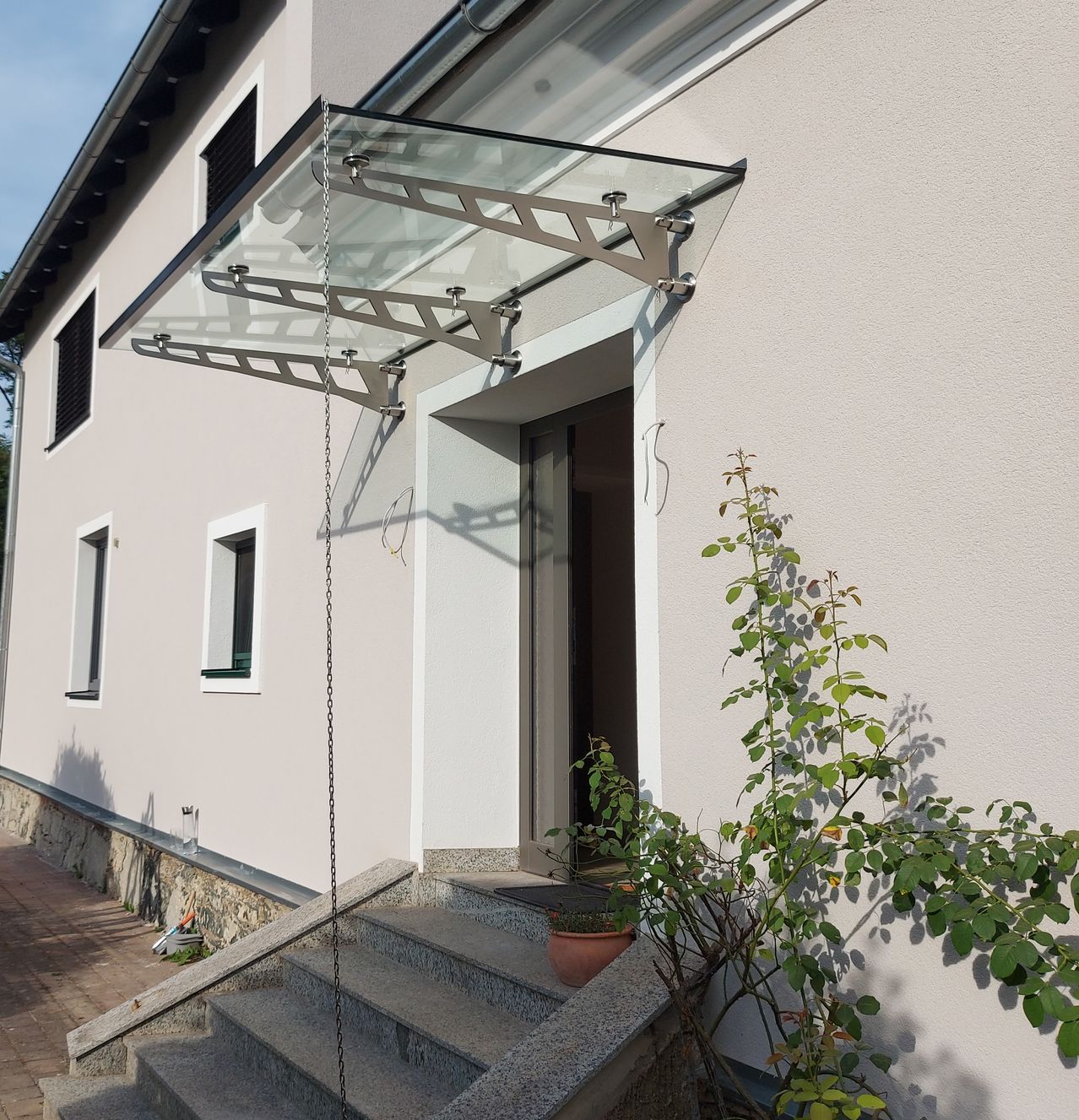 Ein modernes Haus mit einem Glasvordach über dem Eingang, begleitet von einer Topfpflanze und Stufen, die zur Tür führen.