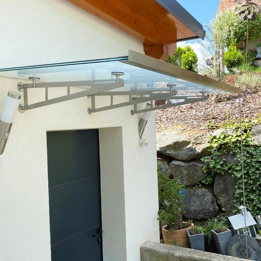 Ein moderner Glasvorbau ist an einer weißen Wand über einer schwarzen Tür befestigt, mit einem geneigten Dach und einem Garten mit Felsen und Pflanzen im Hintergrund.