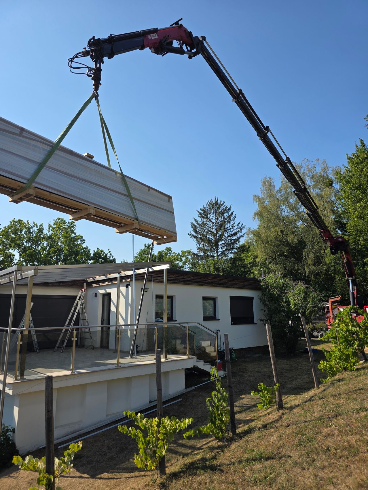 A large wooden beam is being lifted by a crane over a modern house with a glass terrace. The house is surrounded by trees and a vineyard.