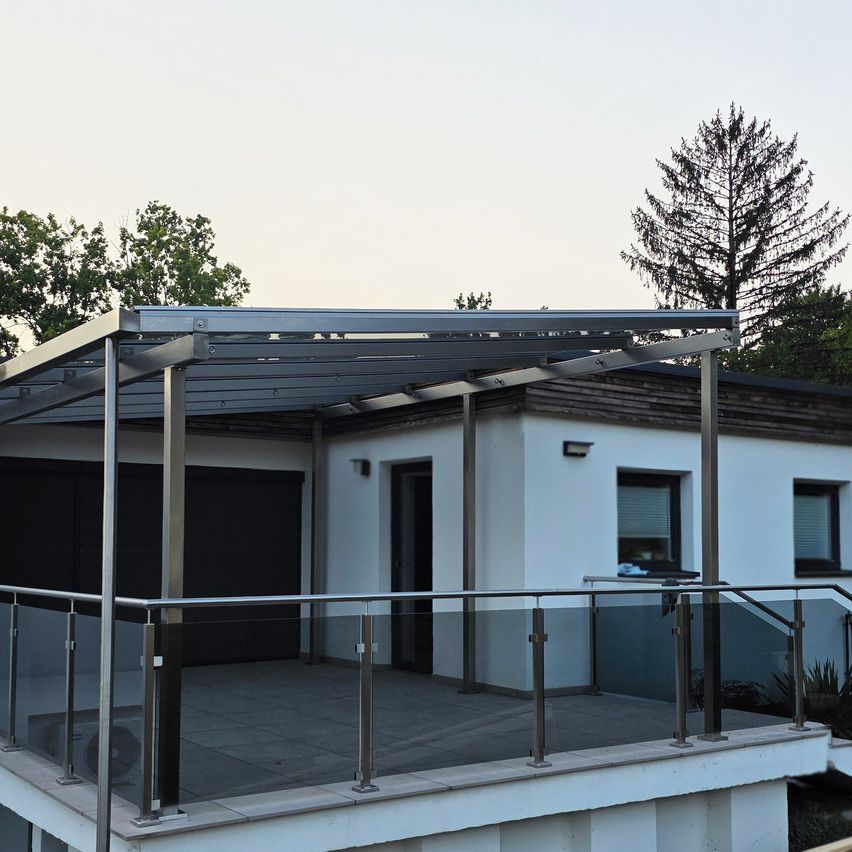 A modern house with a glass-enclosed balcony and a metal pergola roof, surrounded by trees and under a clear sky.