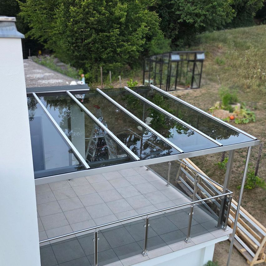An aerial view of a modern balcony with a glass roof and metal railings, surrounded by a grassy area with a greenhouse.