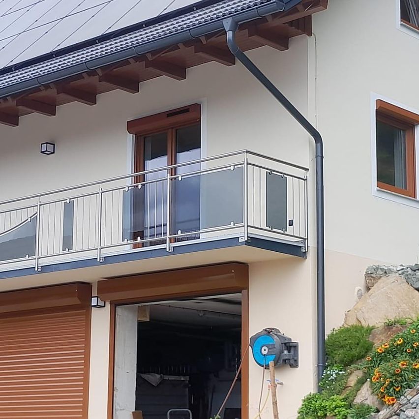 The exterior of a house features a balcony with a glass railing, solar panels on the roof, and a garage entrance with a roll-up door.
