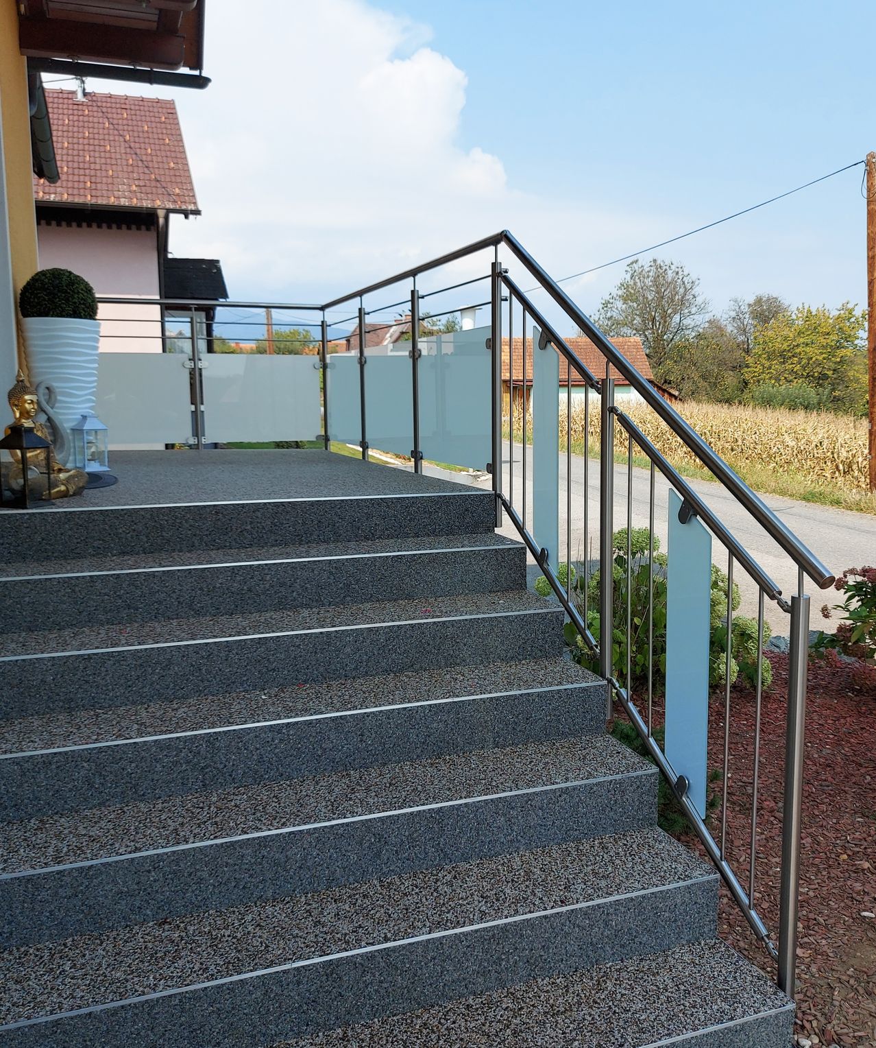 Gray granite steps with metal railings lead to a balcony with a glass fence. A small garden with plants and a statue is in the foreground.