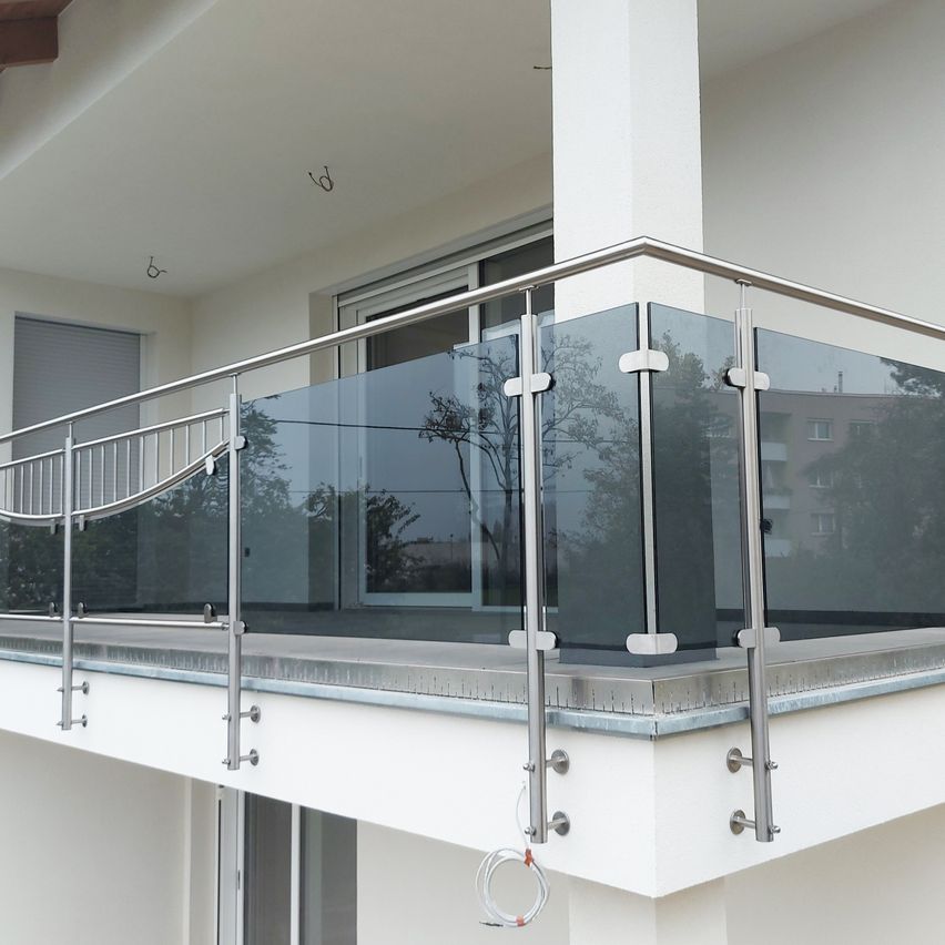 A modern balcony with a glass railing and metal supports, featuring a white wall and a glass panel reflecting a tree and building.