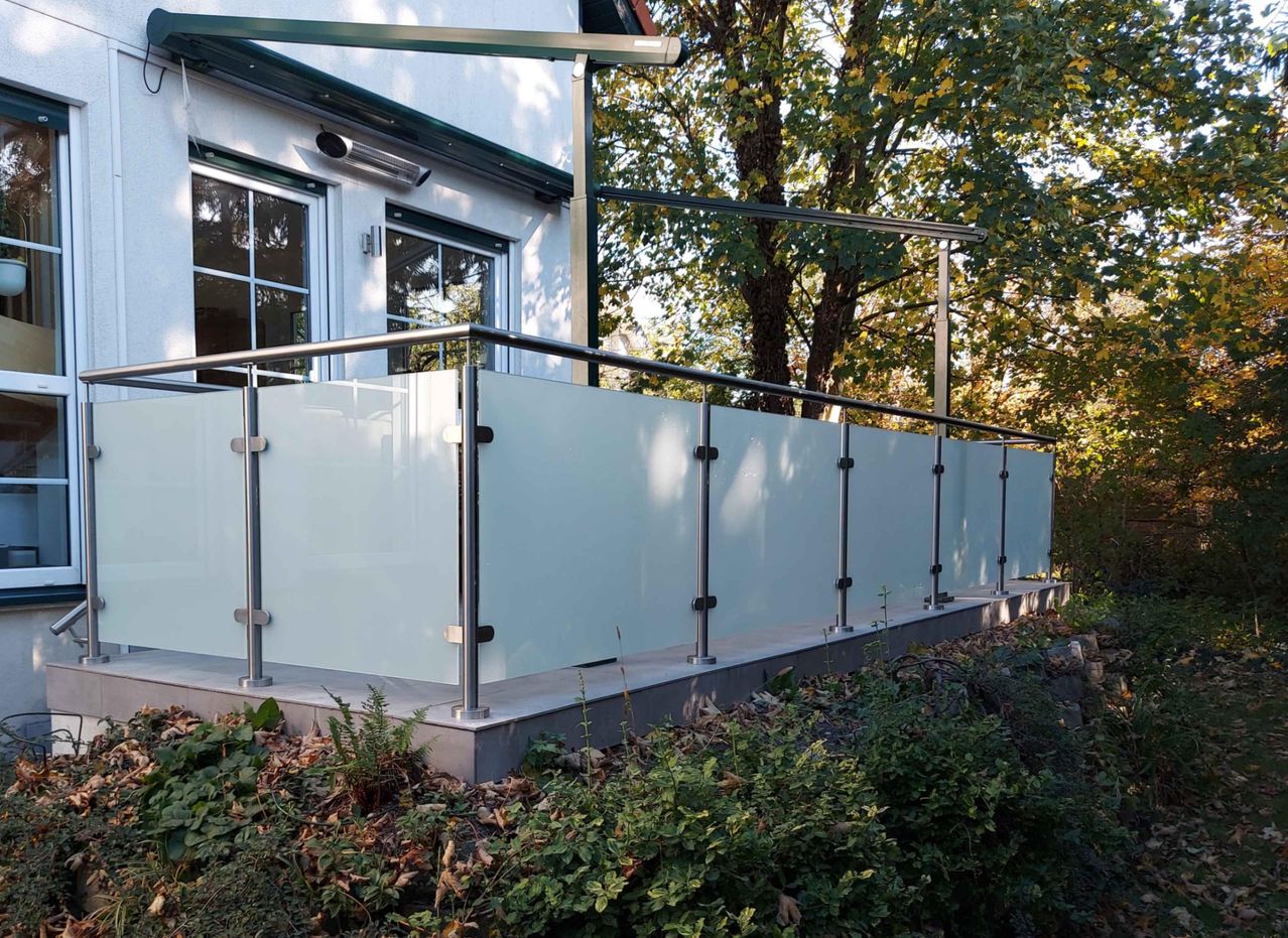 Ein modernes Haus mit einem Balkon, Glasgeländern und einem Vordach. Der Balkon hat eine weiße Wand und Metallgeländer. Ein Baum steht hinter dem Balkon.