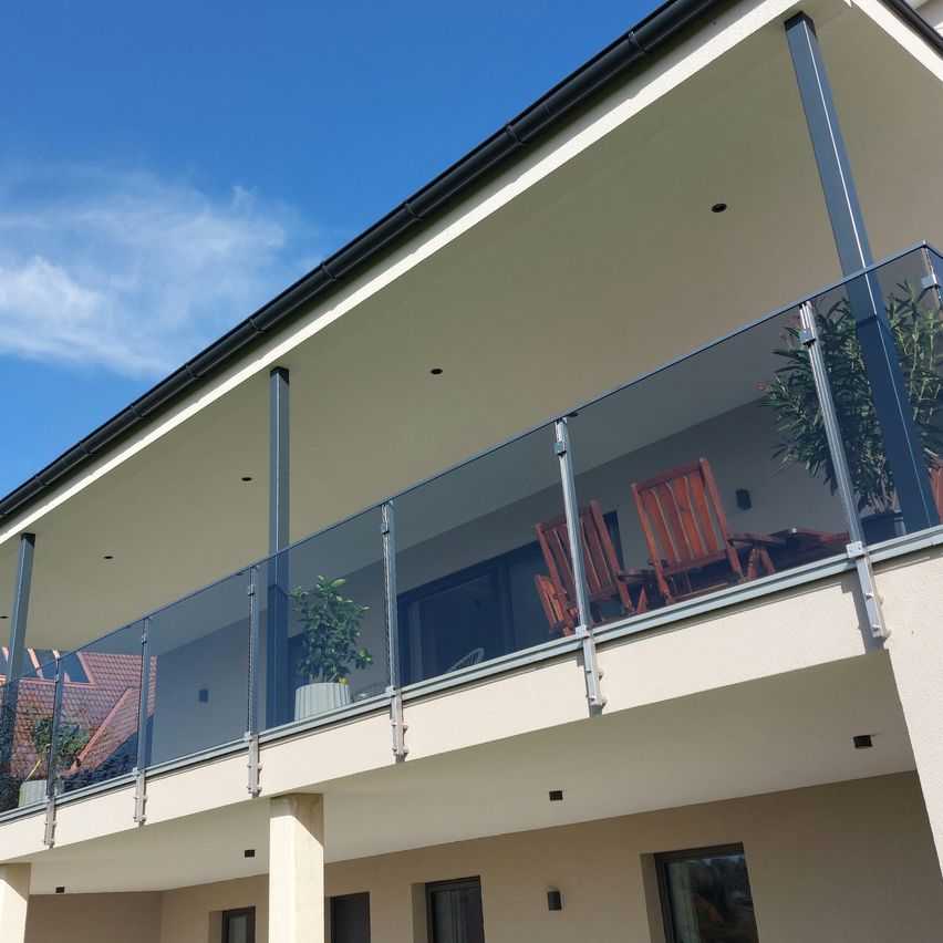 Ein modernes Apartmentgebäude mit einem Glasbalkon, ausgestattet mit Holzstühlen und Topfpflanzen. Der Balkon liegt unter einem klaren blauen Himmel.